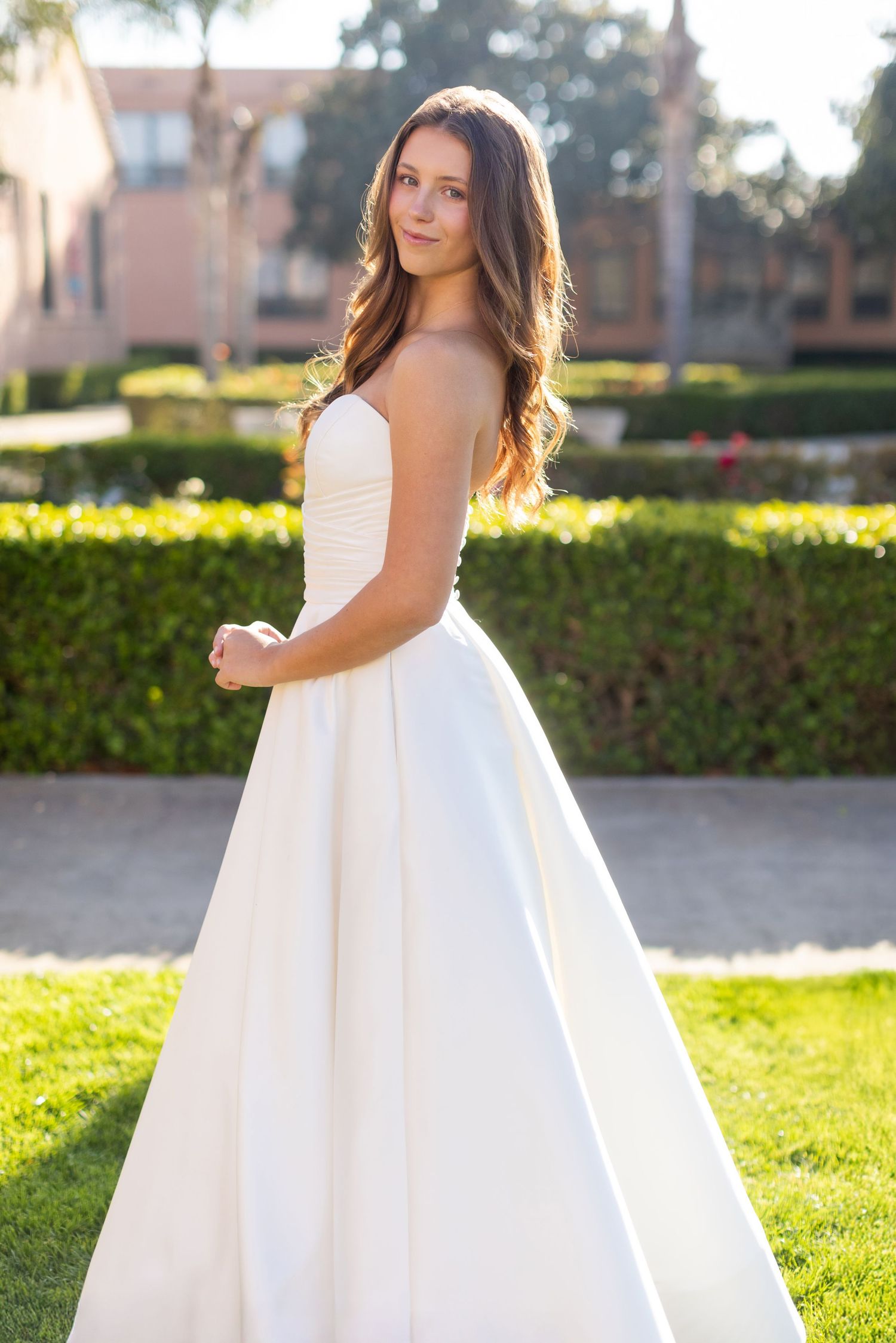 Long white strapless satin ball gown wedding dress photographed outdoors in sunlit garden setting.