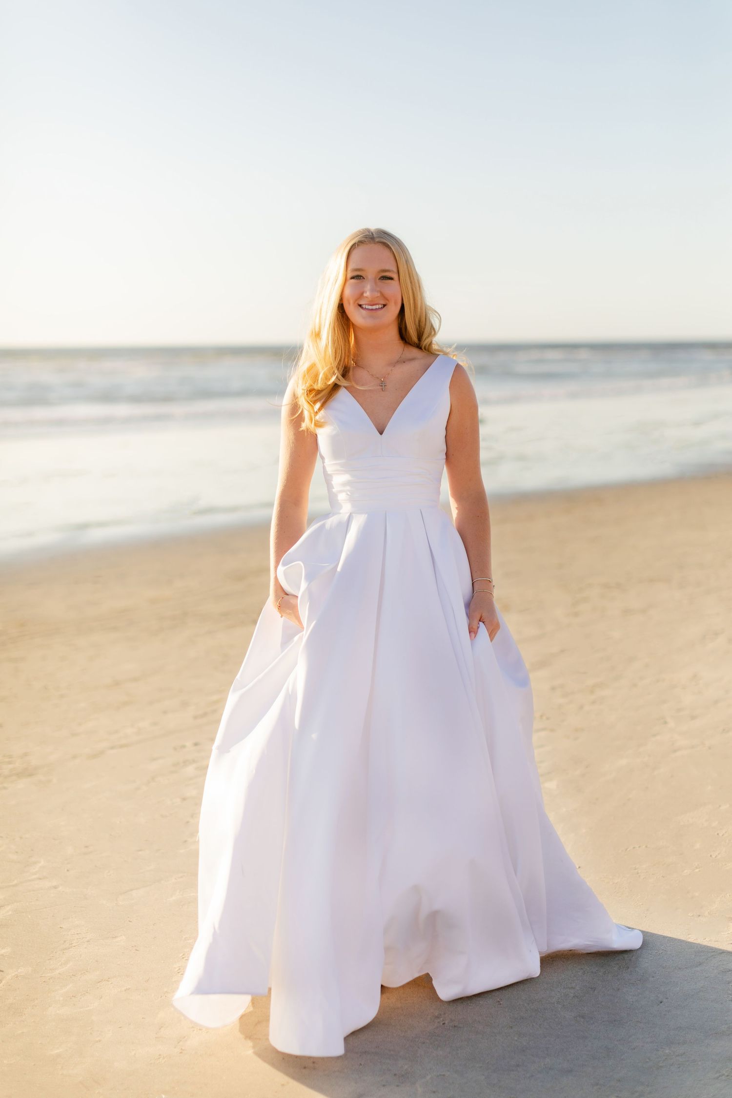 A woman in an elegant white wedding dress poses on a sunny beach at golden hour.