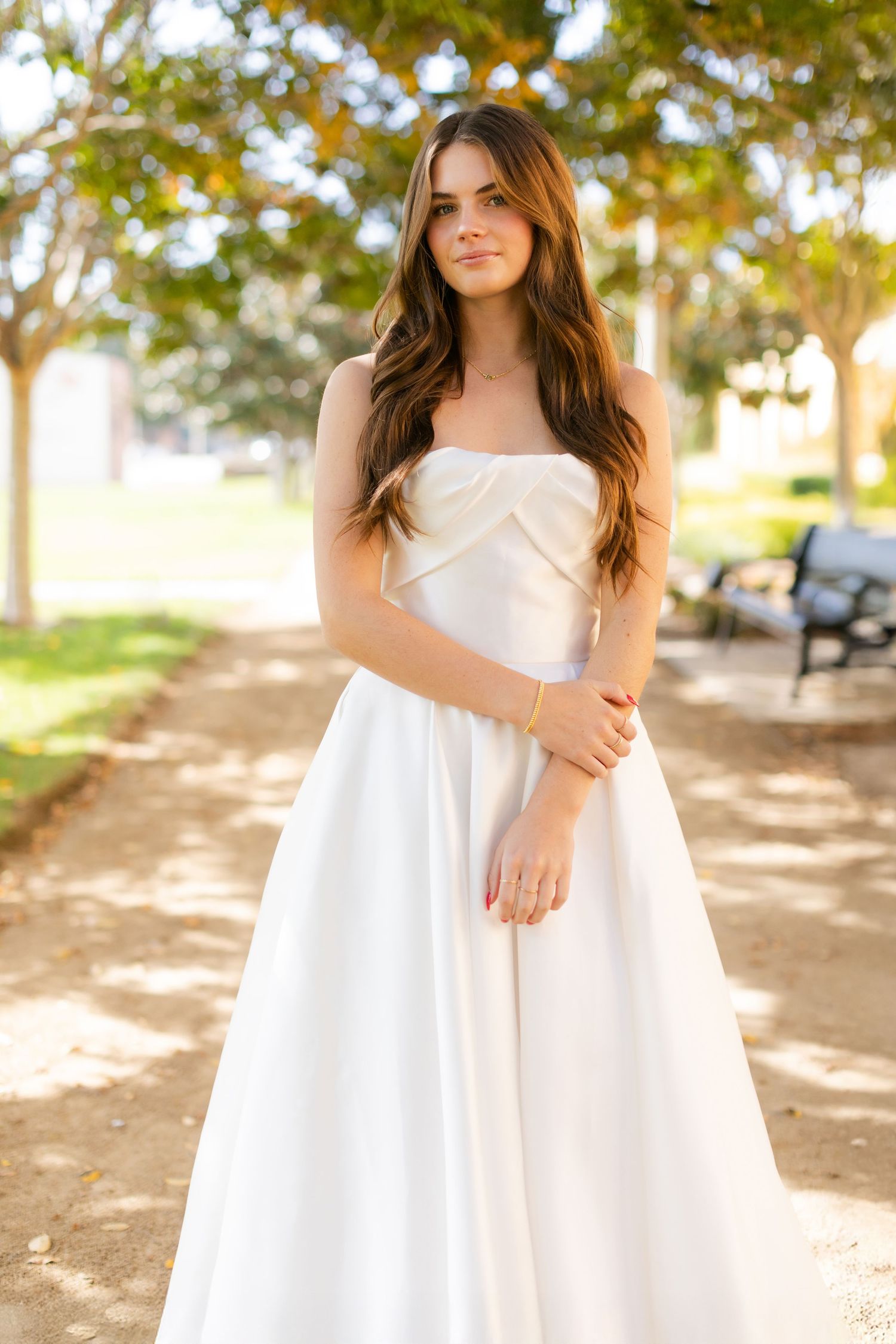 Beautiful strapless white ball gown dress with a fitted bodice and flowing skirt shown outdoors in a sunlit park setting.