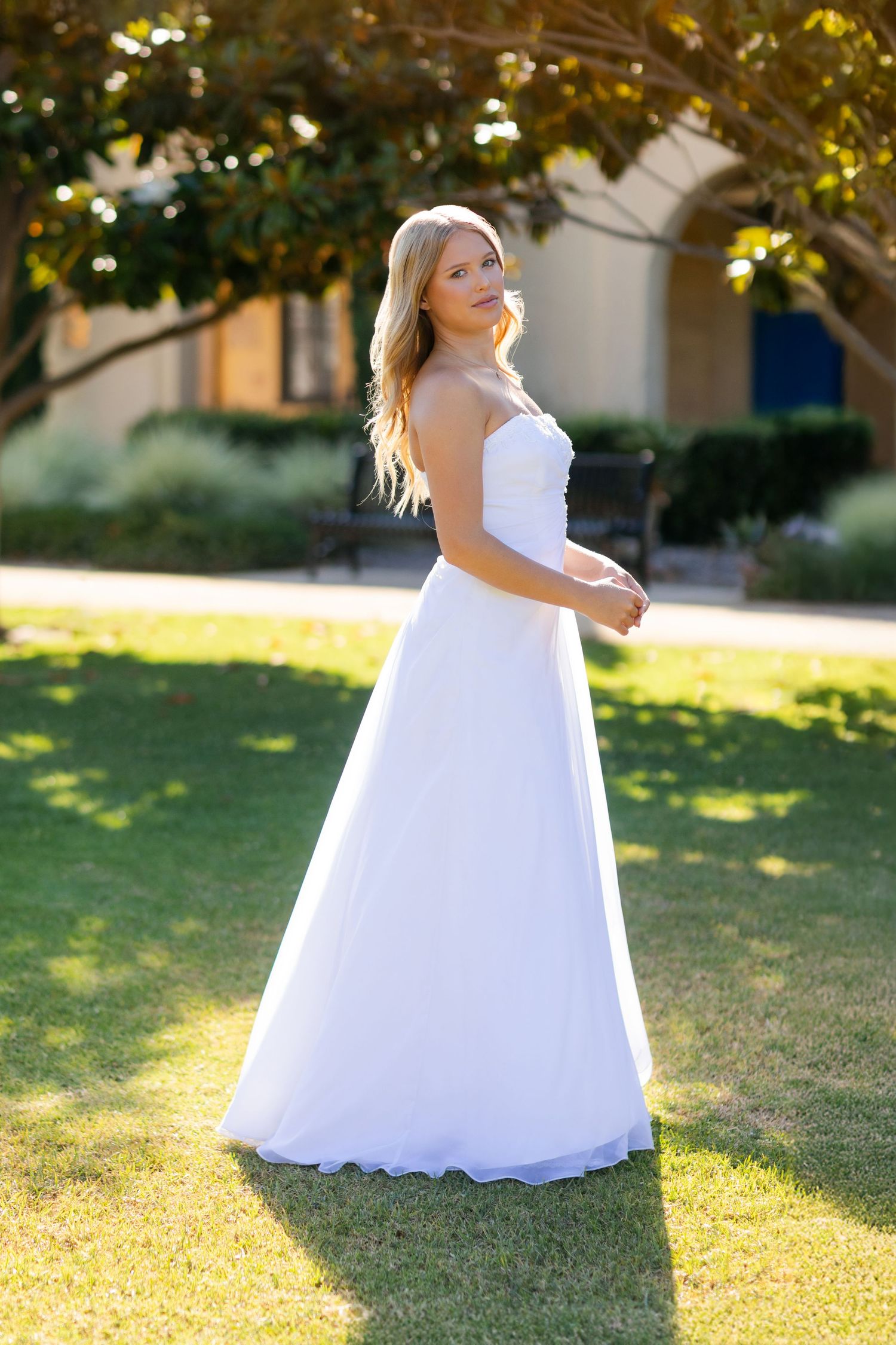 A person in a flowing white wedding gown stands on a sunlit lawn in front of a building during golden hour.