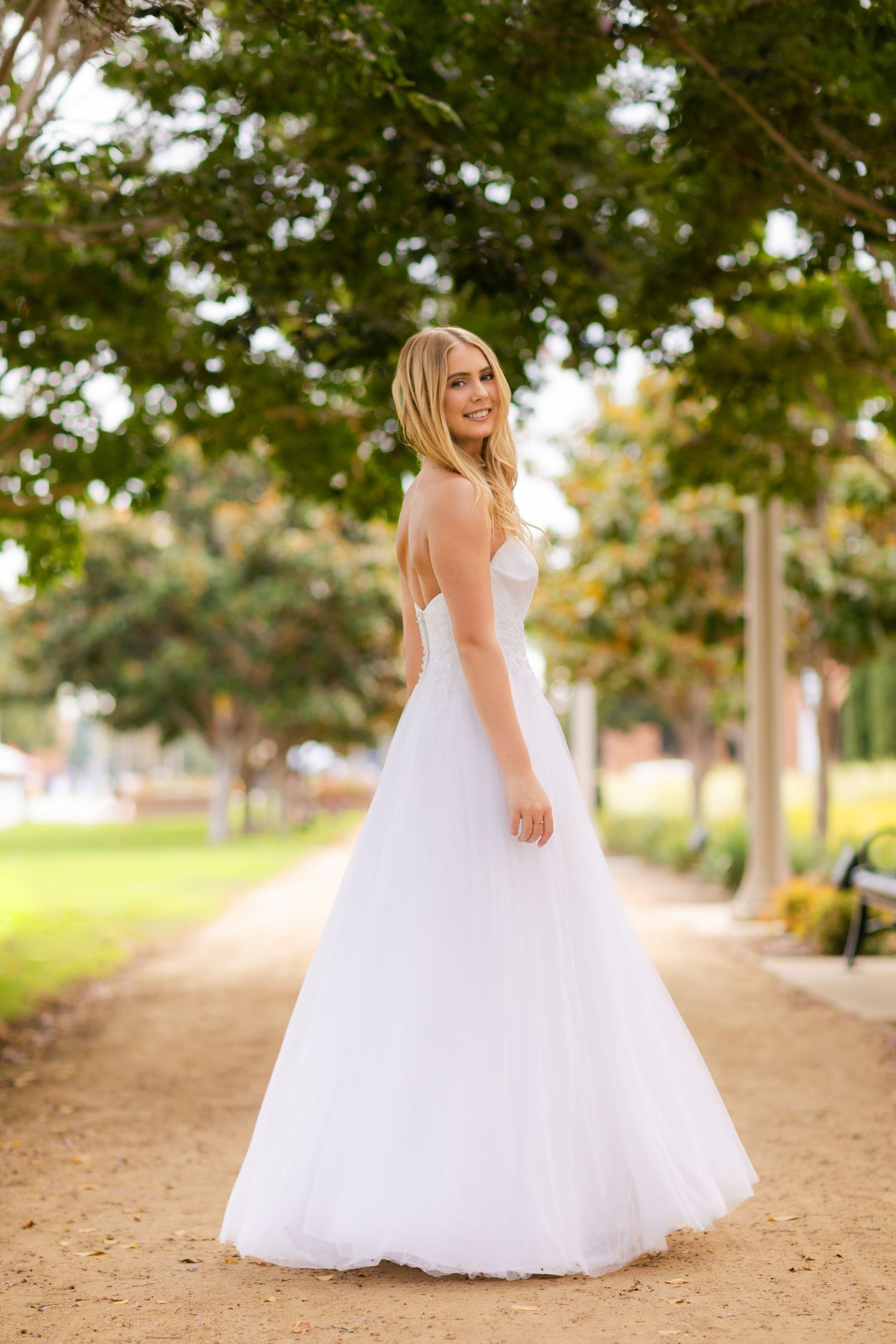 A person in an elegant white wedding gown stands on a tree-lined dirt path during golden hour.