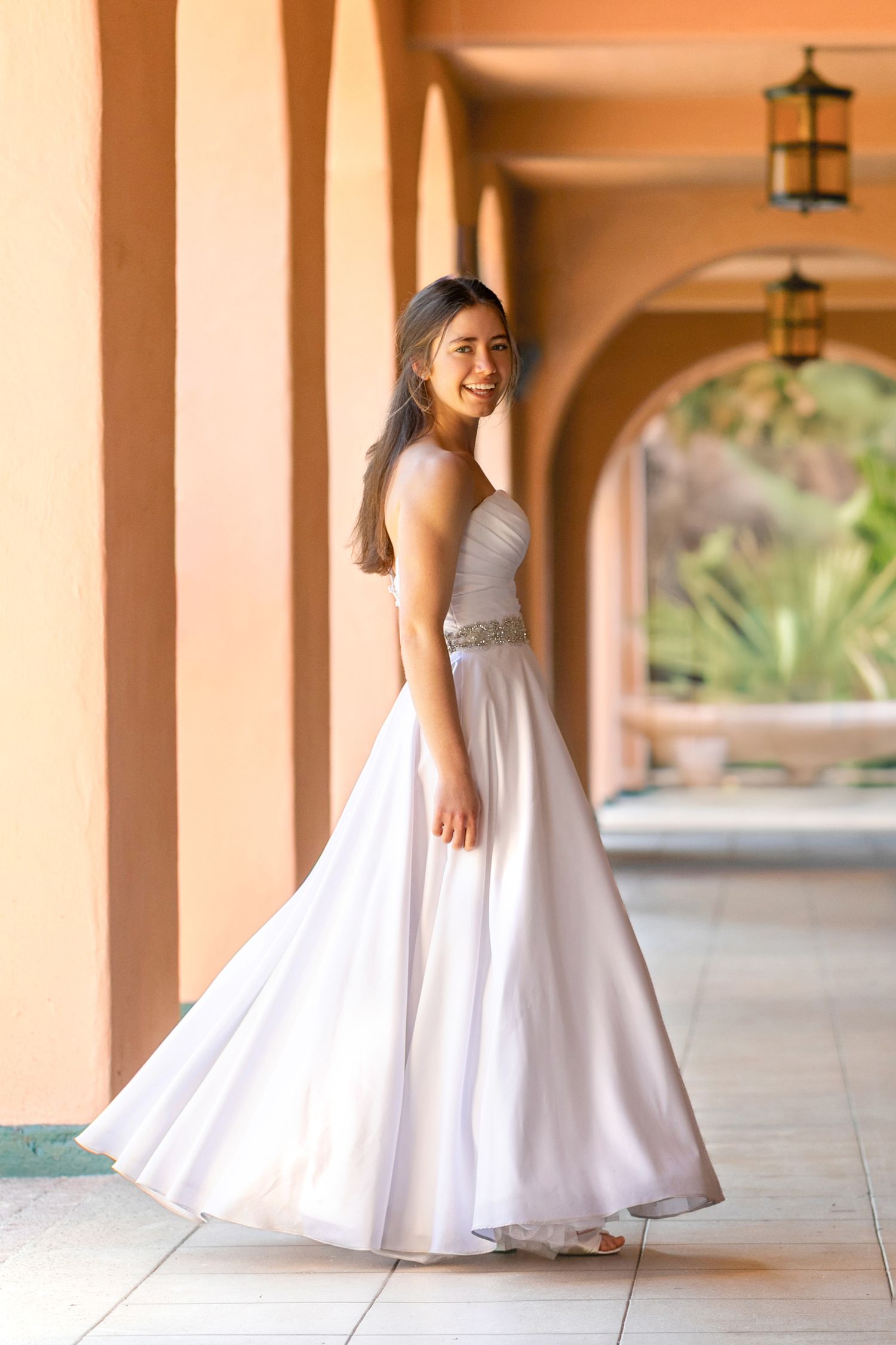 A woman in an elegant white formal gown poses gracefully under an arched walkway with peach-colored walls and lanterns.