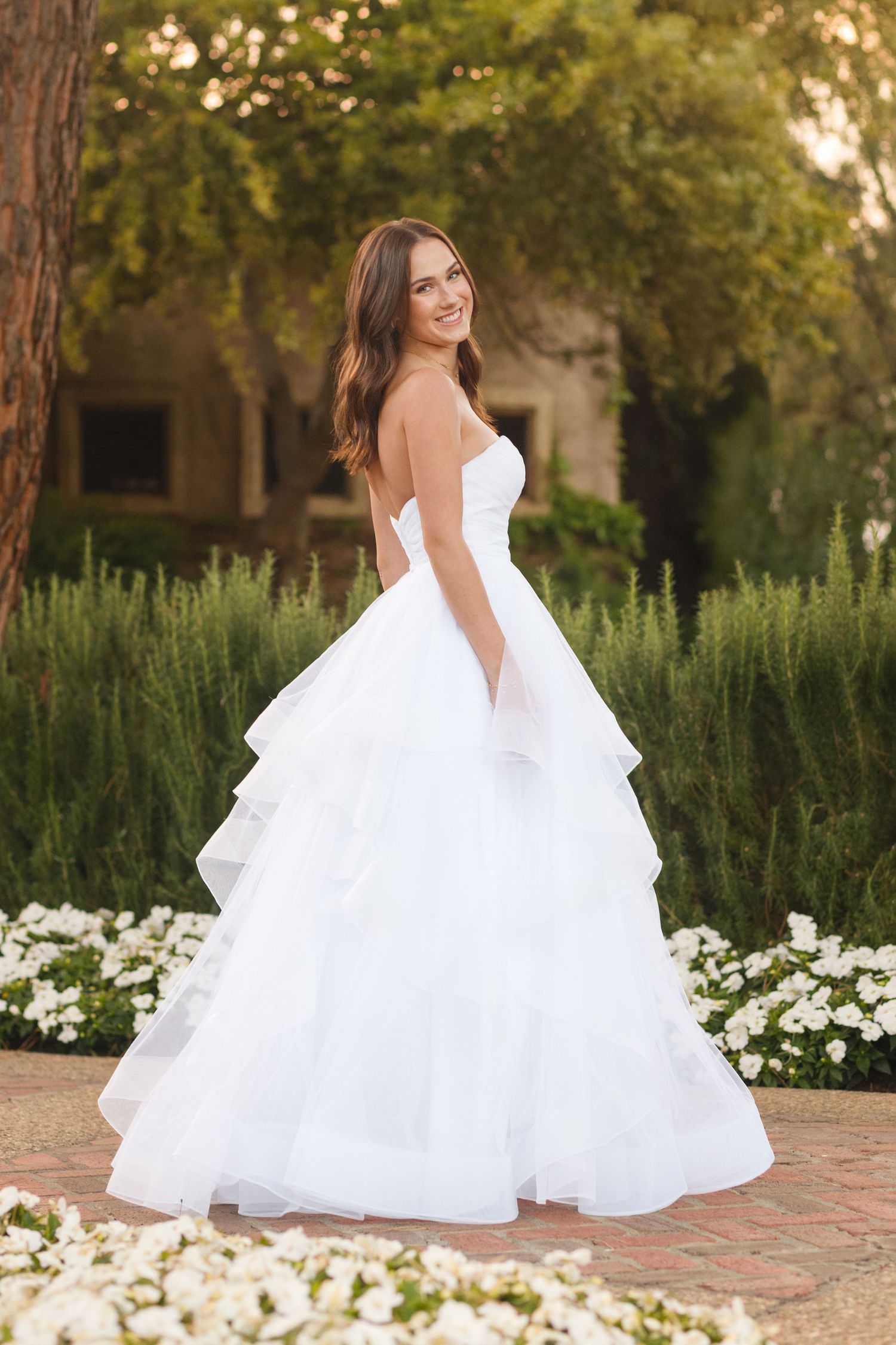 A strapless white wedding gown with tiered skirt and sweetheart neckline photographed in a garden with white flowers.