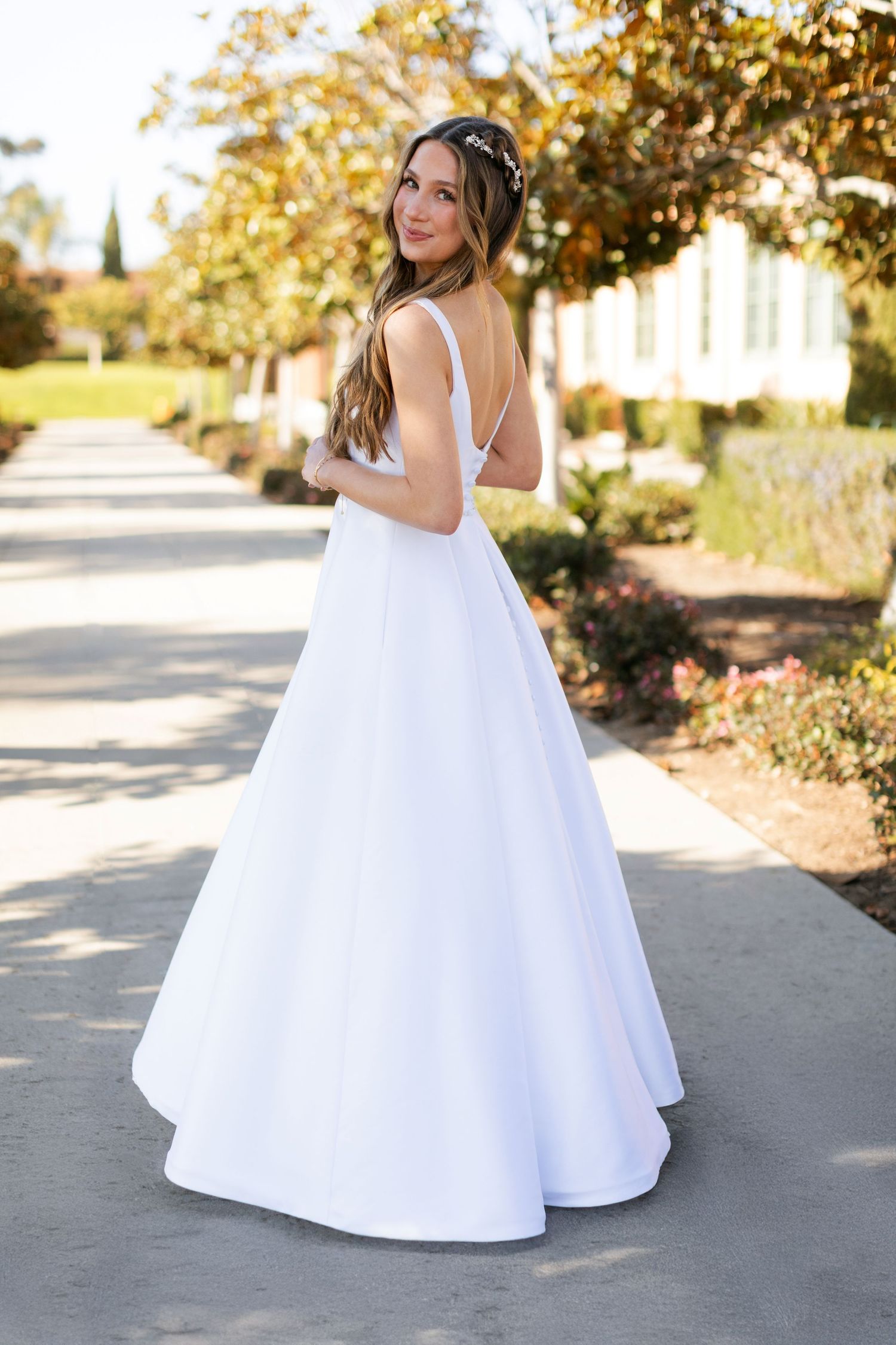 A bride in a flowing white wedding gown poses outdoors on a path surrounded by autumn foliage and gardens.