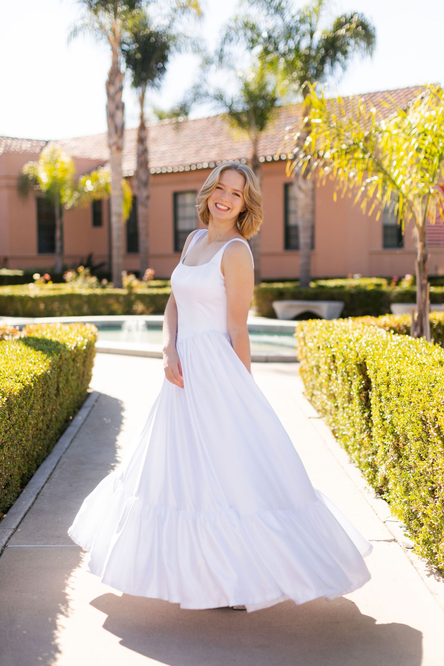 A white sundress flowing in the sunlight along a garden path with palm trees and manicured hedges in the background.