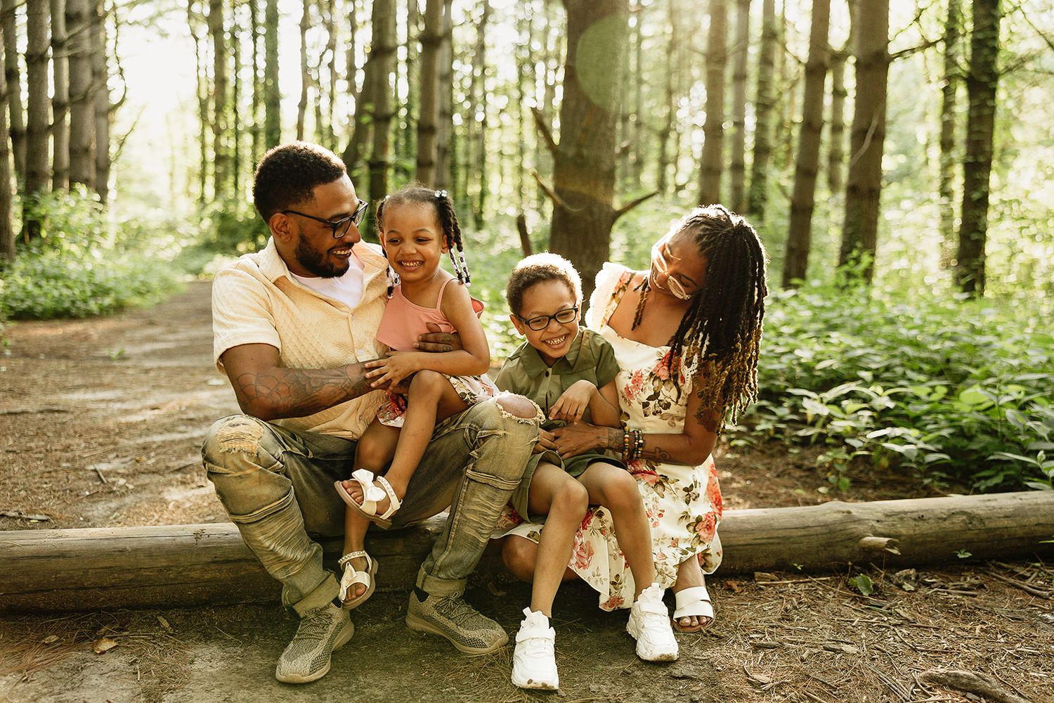 A family of four sits together on a fallen log in a sunlit forest sharing a joyful moment.