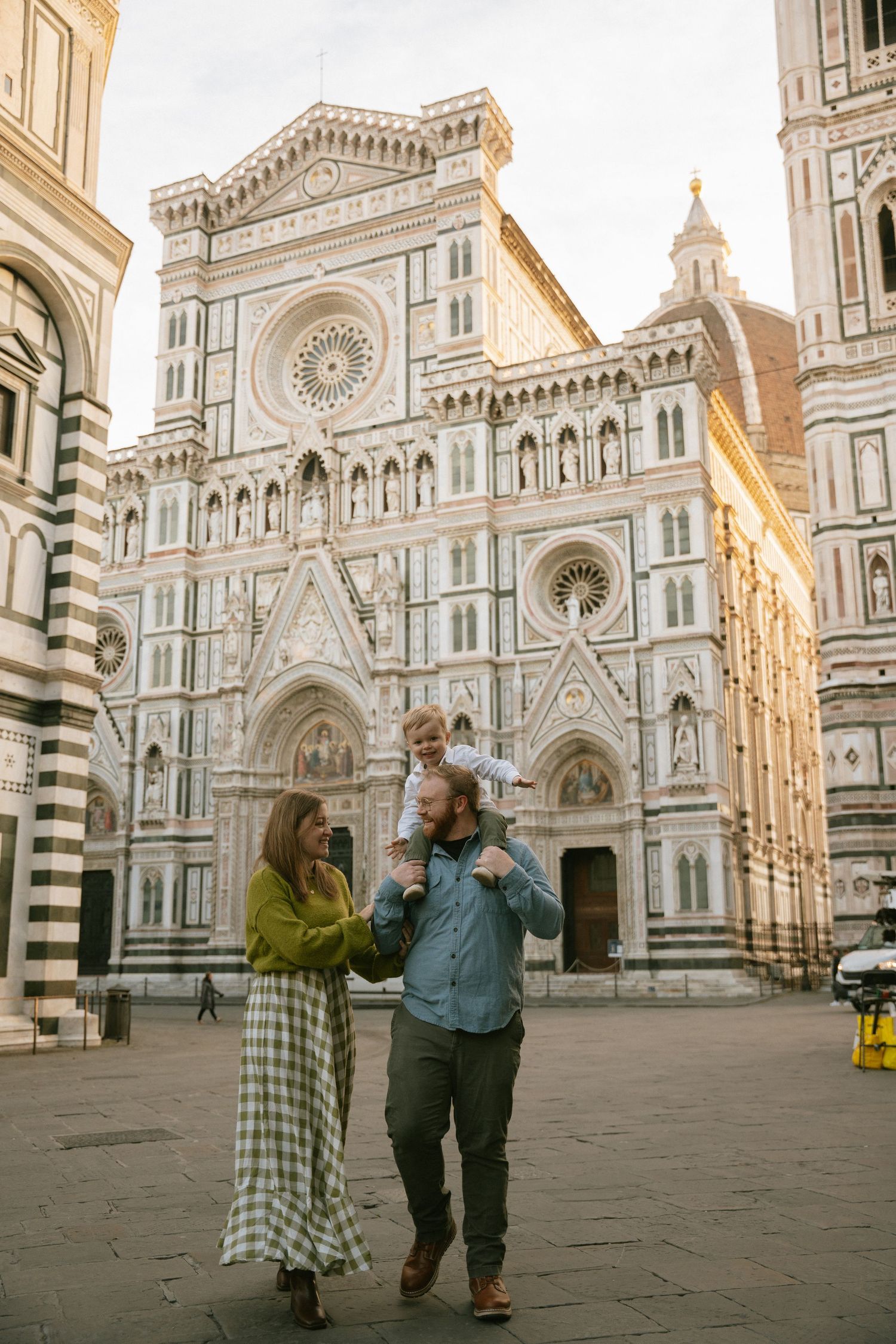 Family photo of a couple walking with their young child on the man's shoulders in front of the Florence Cathedral in Italy.