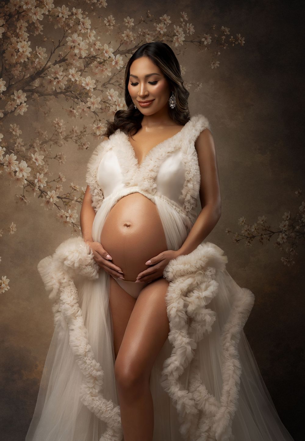Pregnant woman in white fluffy robe maternity gown holding belly with floral backdrop.