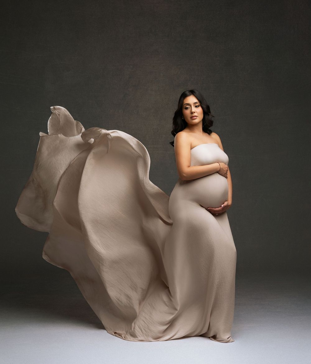Pregnant woman in flowing beige strapless gown with fabric billowing dramatically against dark studio background.