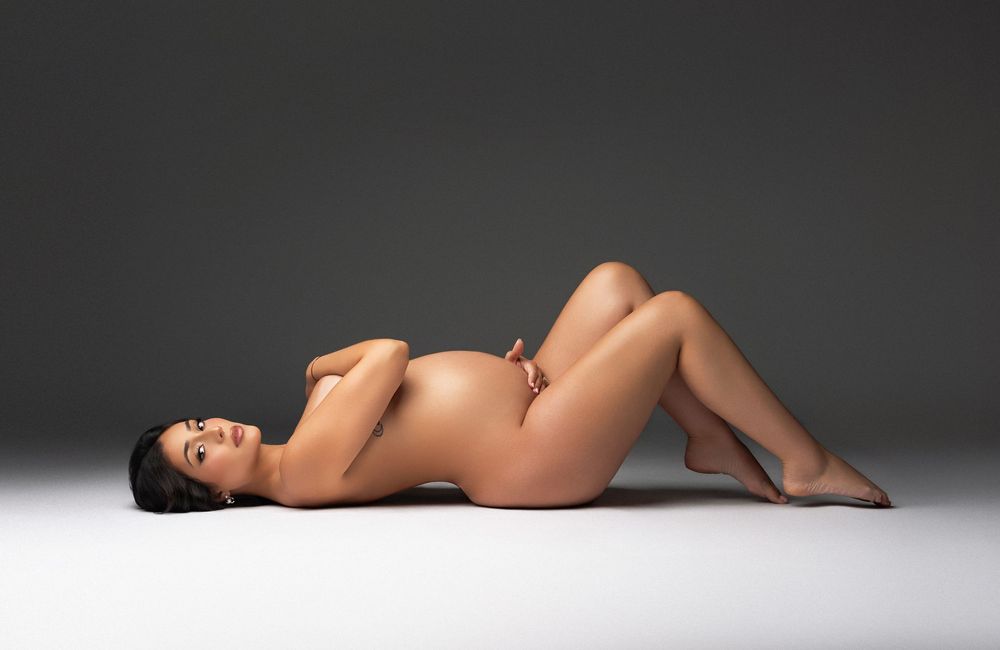 A nude pregnant woman reclines gracefully on a studio floor against a gray background, showcasing her baby bump.