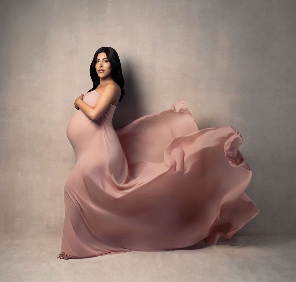 Pregnant woman in flowing pink maternity gown poses gracefully against neutral studio backdrop.