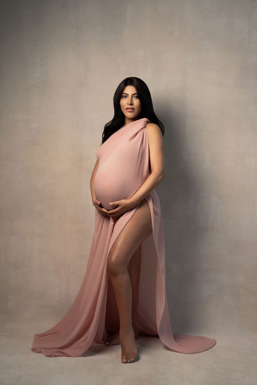 Pregnant woman draped in flowing pink chiffon fabric, cradling her baby bump in an elegant maternity studio portrait.