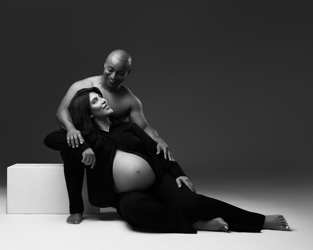 Black and white maternity photo of a pregnant woman in black outfit with her bald partner in a studio setting.