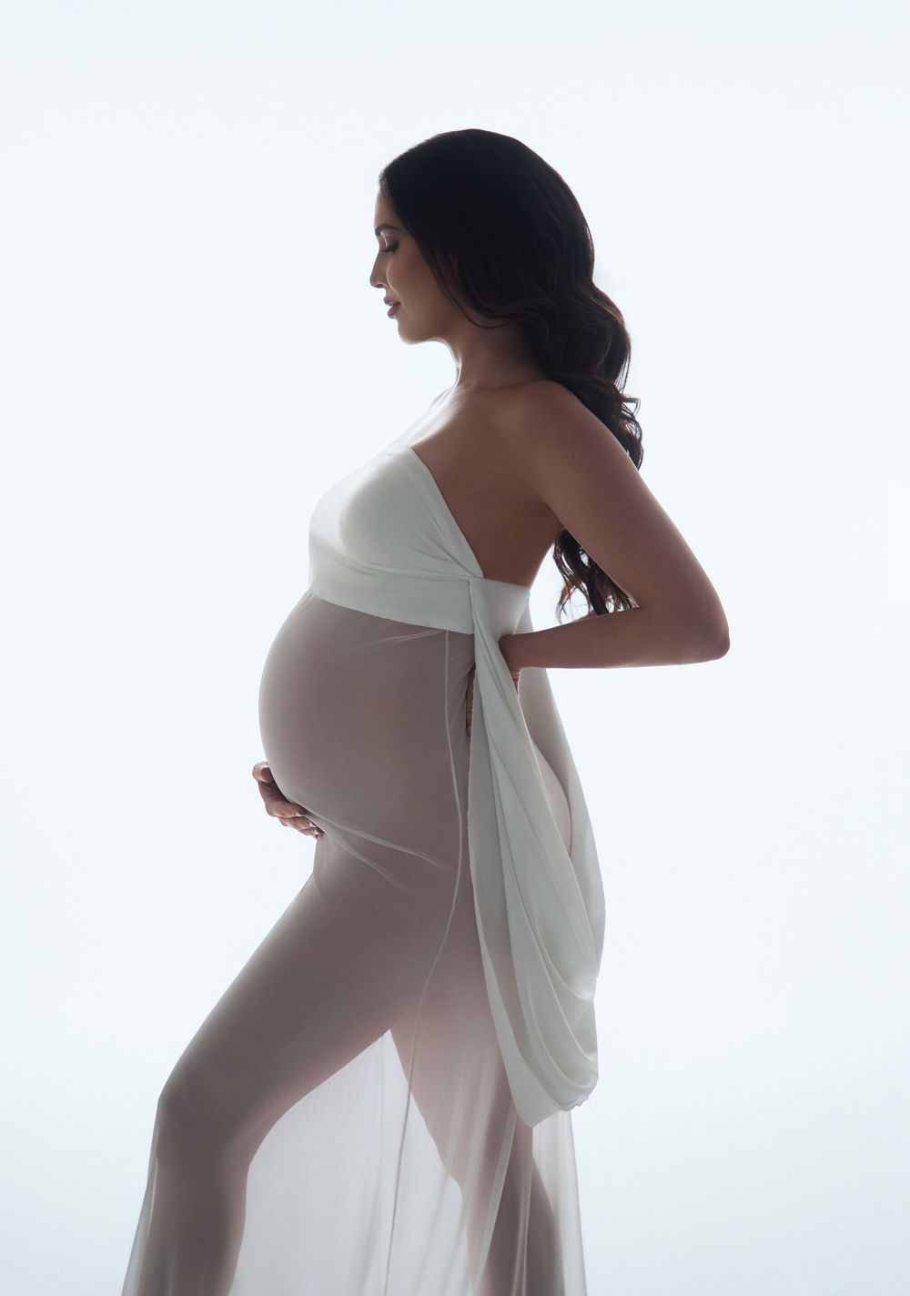 Elegant pregnant woman in sheer white draped fabric poses sideways, showcasing baby bump in a studio maternity portrait.