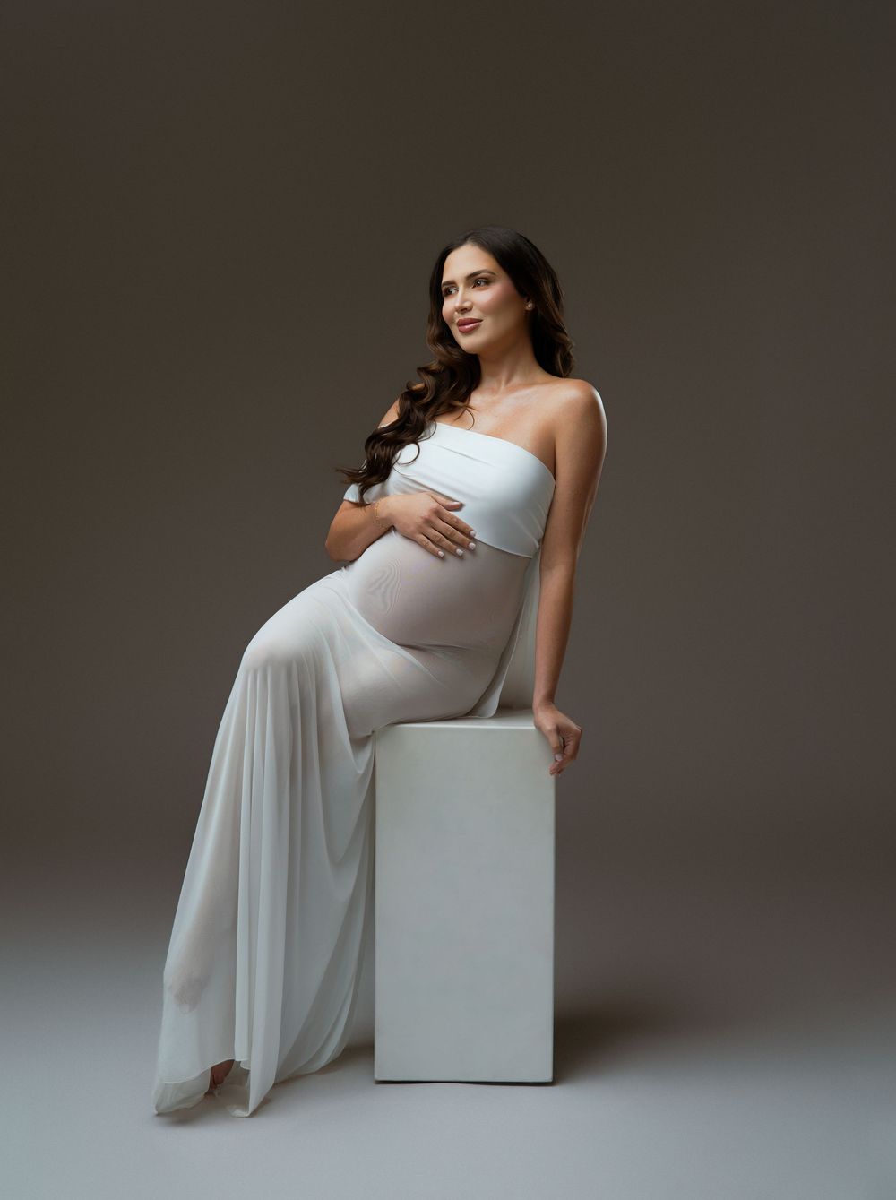 Pregnant woman in white strapless gown sitting on white cube, cradling baby bump in professional maternity photo.