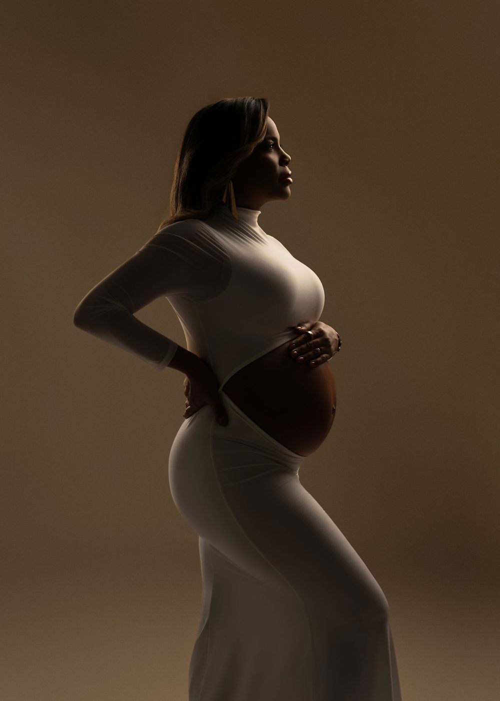 Elegant pregnant woman in white dress cradling baby bump in dramatic studio lighting silhouette.