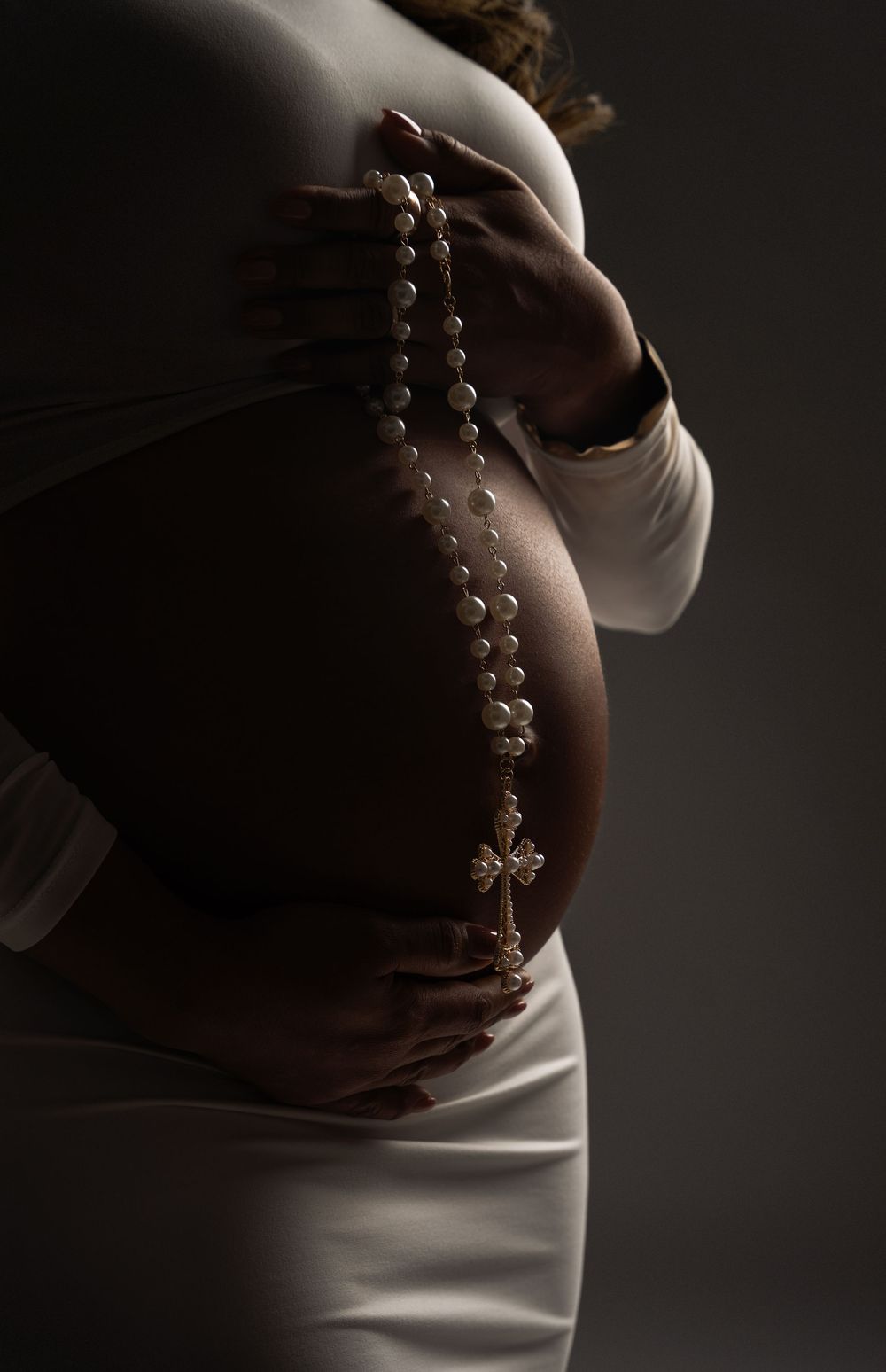 Pregnant woman in white dress holding rosary beads with cross over baby bump in dramatic studio lighting.
