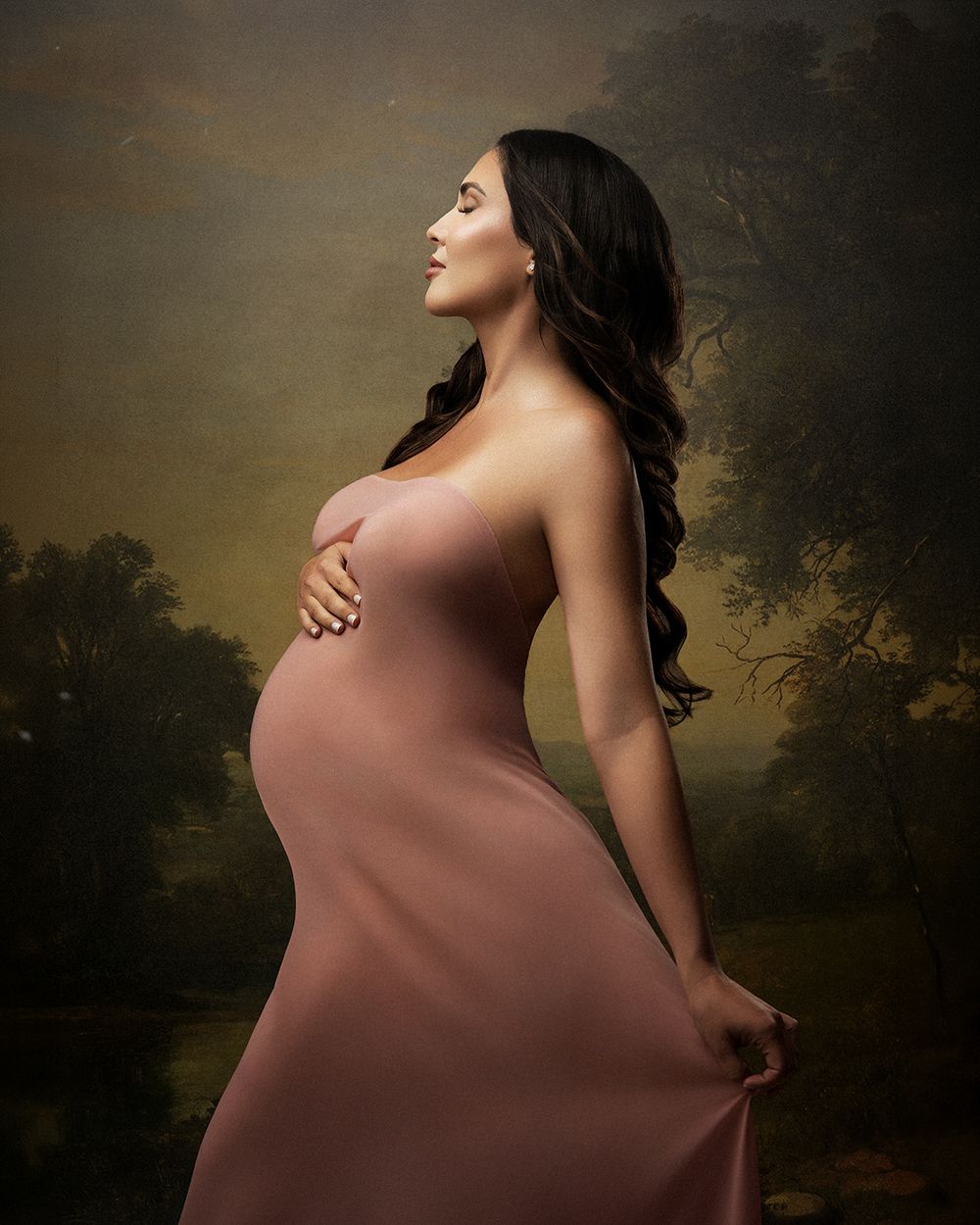 Pregnant woman in pink strapless gown holding belly, posing against a classic painterly landscape backdrop.
