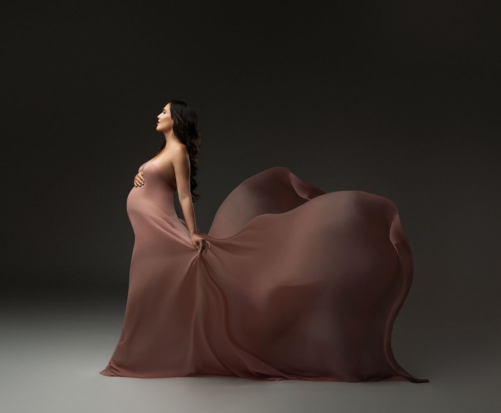 Pregnant woman in flowing mauve gown with dramatic fabric train poses gracefully against dark studio background.