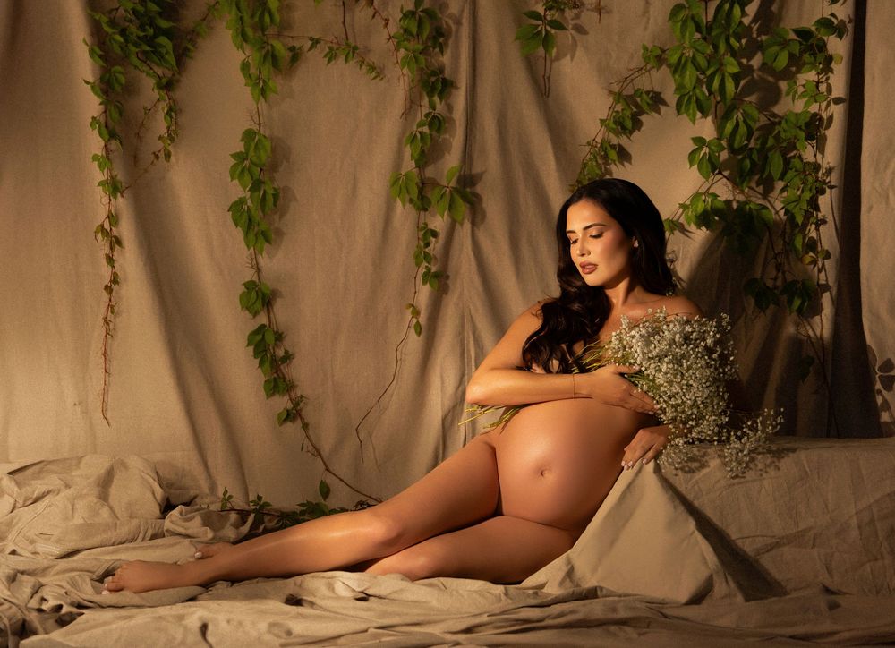 Pregnant woman in elegant maternity photo shoot, reclining on draped fabric with ivy backdrop in warm studio lighting.