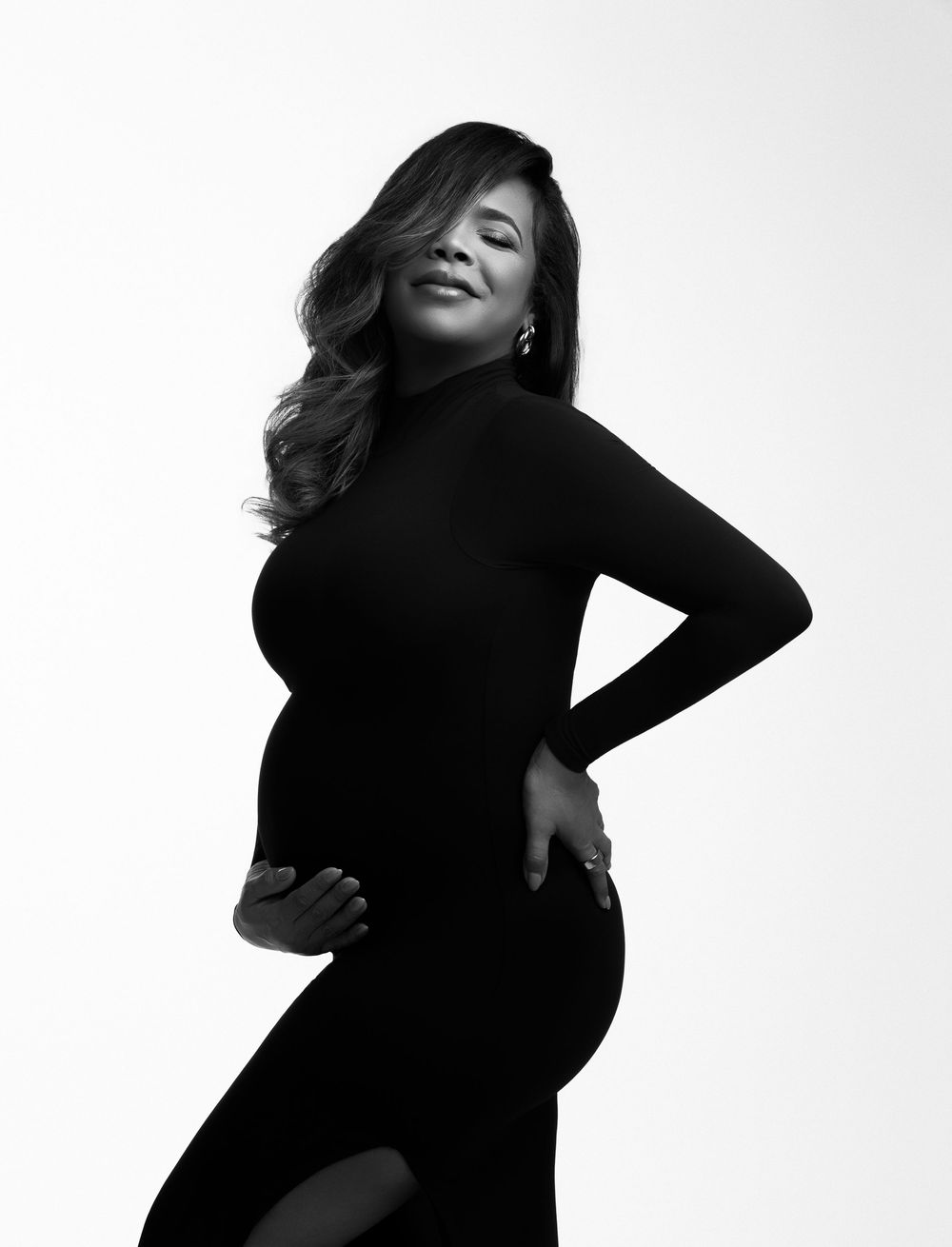 Black and white maternity portrait of a woman in a black turtleneck dress, elegantly posing with hand on hip.