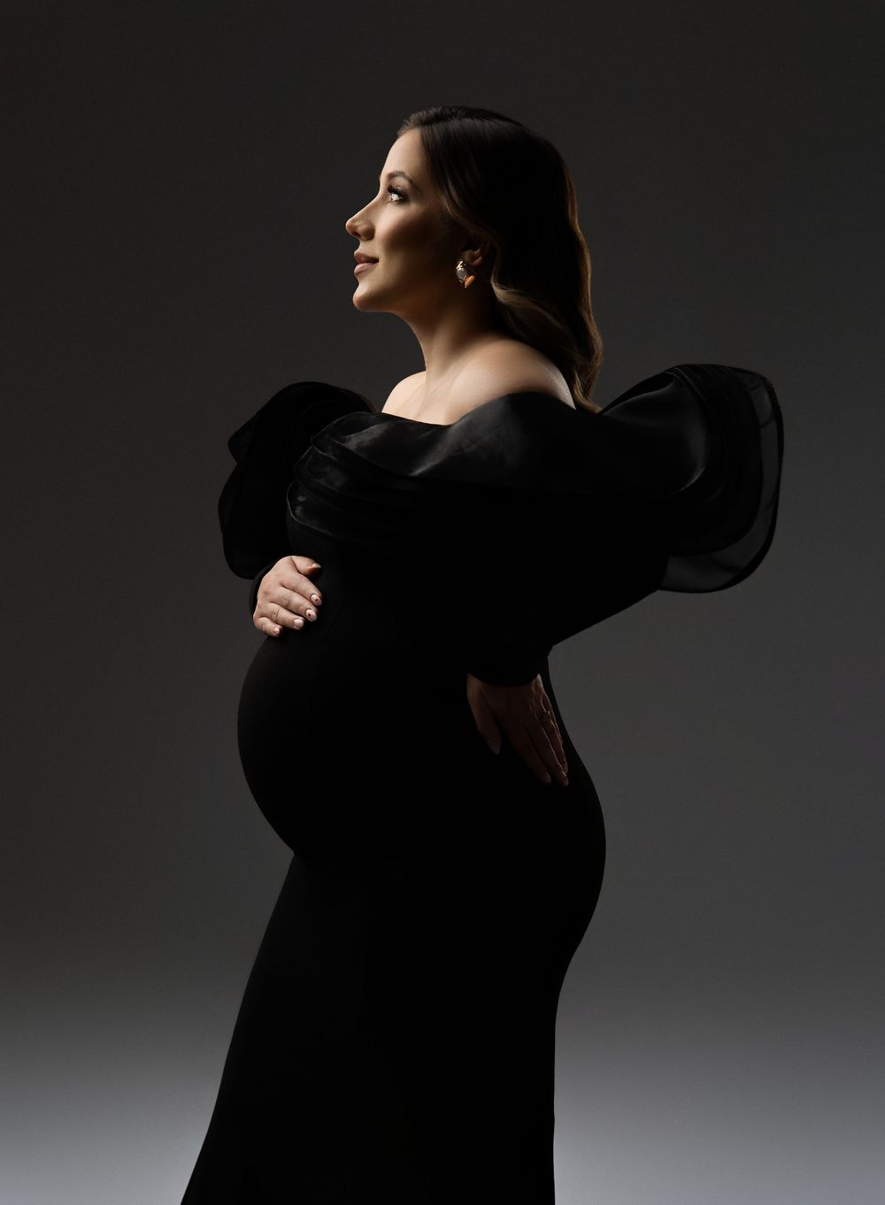 Silhouetted figure in black clothing performs expressive dance movements against dark gray background in dramatic lighting.
