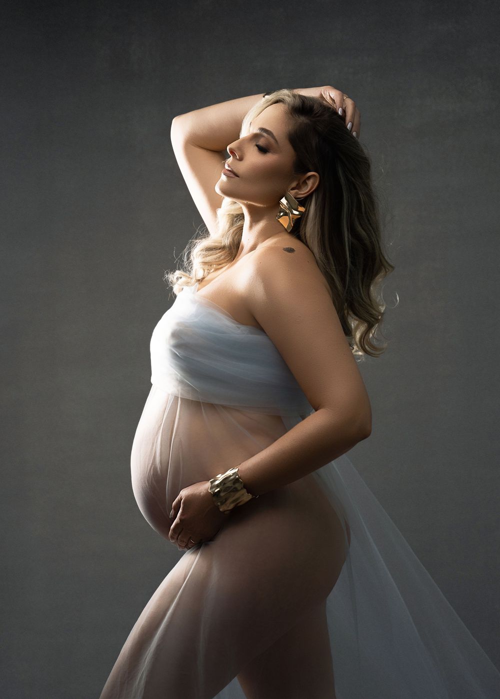Elegant maternity portrait in soft white fabric against dark background with dramatic lighting and ethereal mood.