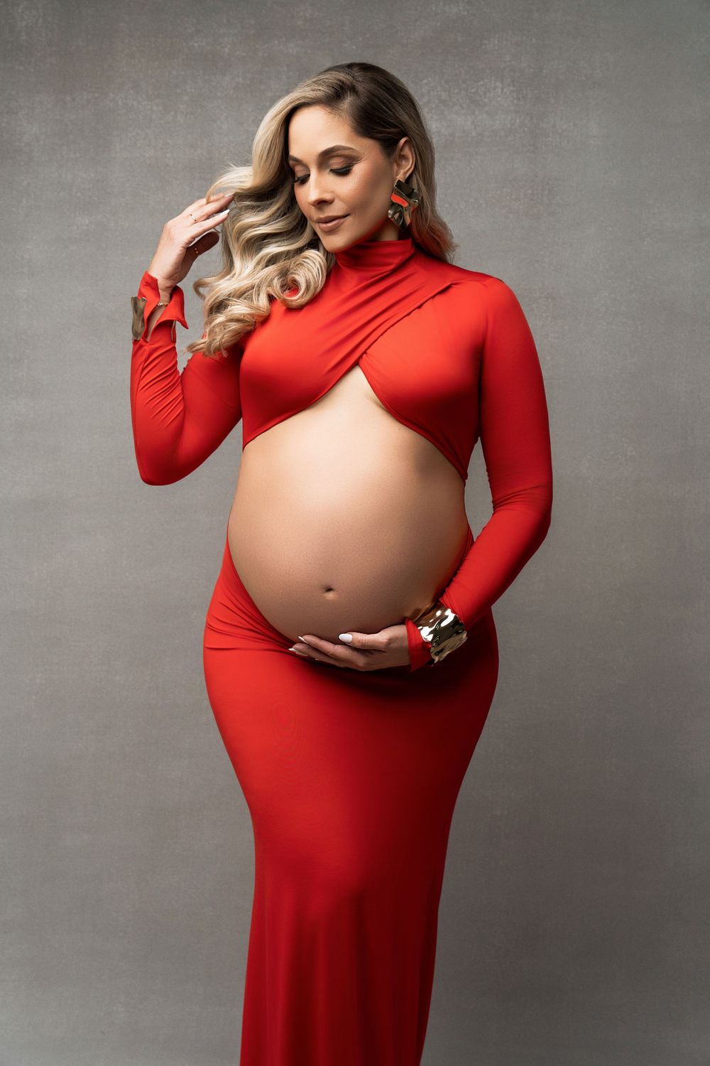 A pregnant woman in a fitted red long sleeve dress poses elegantly against a gray studio backdrop.
