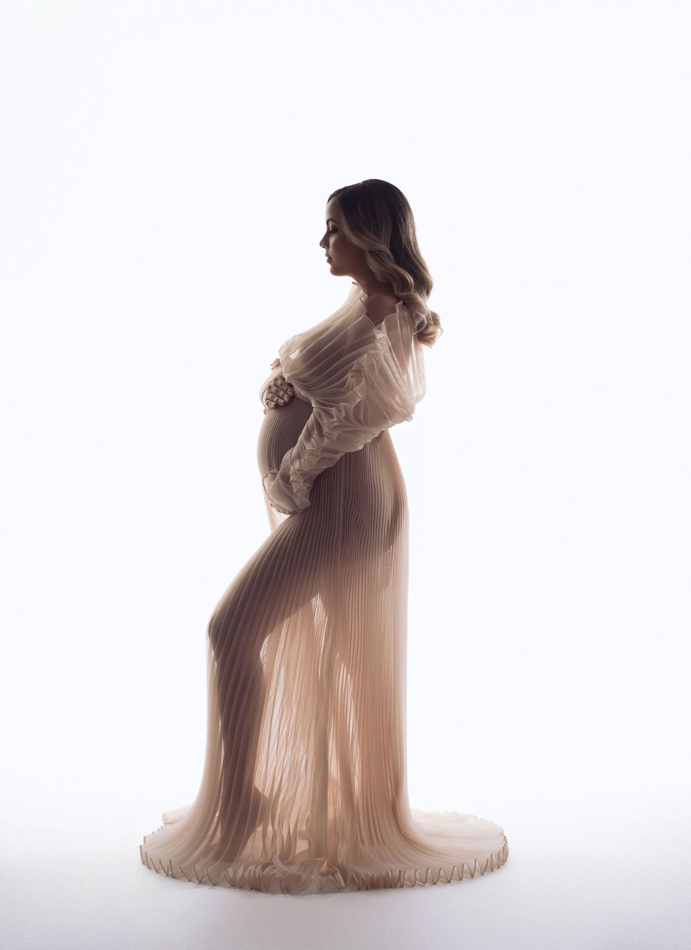 Pregnant woman in elegant beige pleated maternity gown posing sideways against white background in professional studio photo.