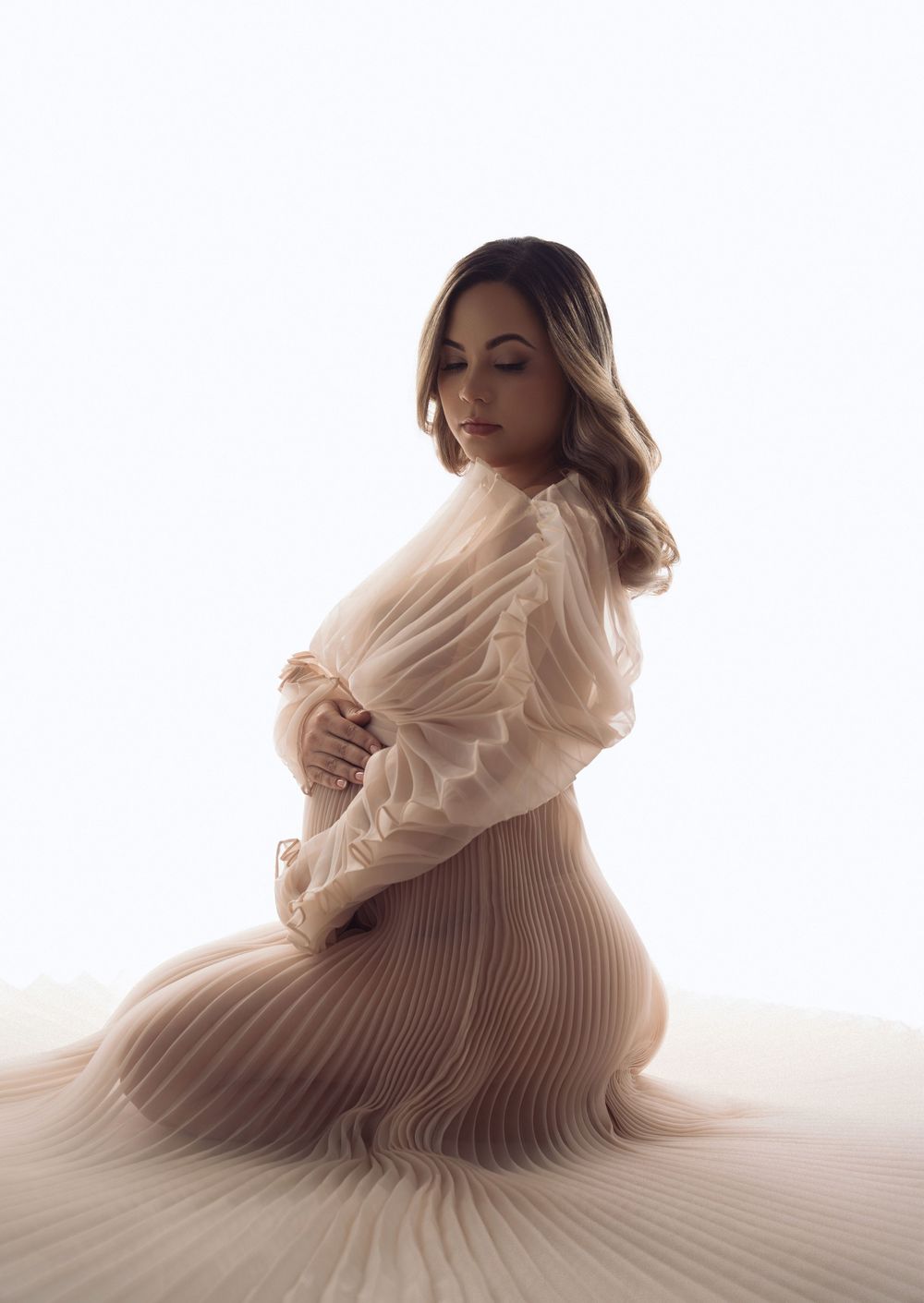 Pregnant woman in elegant cream pleated dress kneeling gracefully against white background.