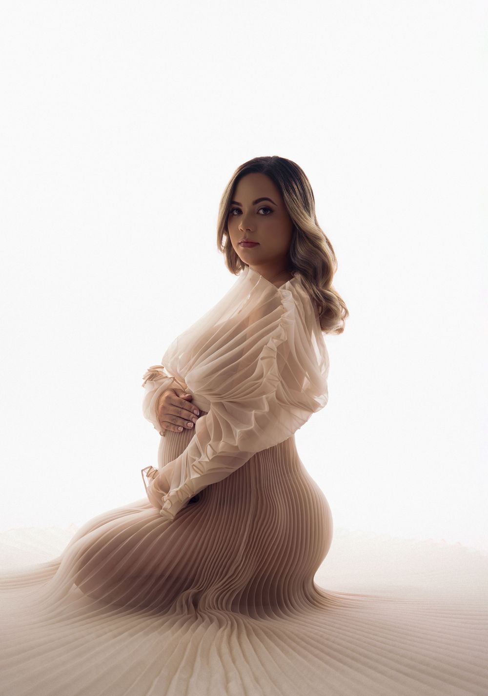 Expectant mother in sheer cream pleated gown posing gracefully on white studio backdrop.
