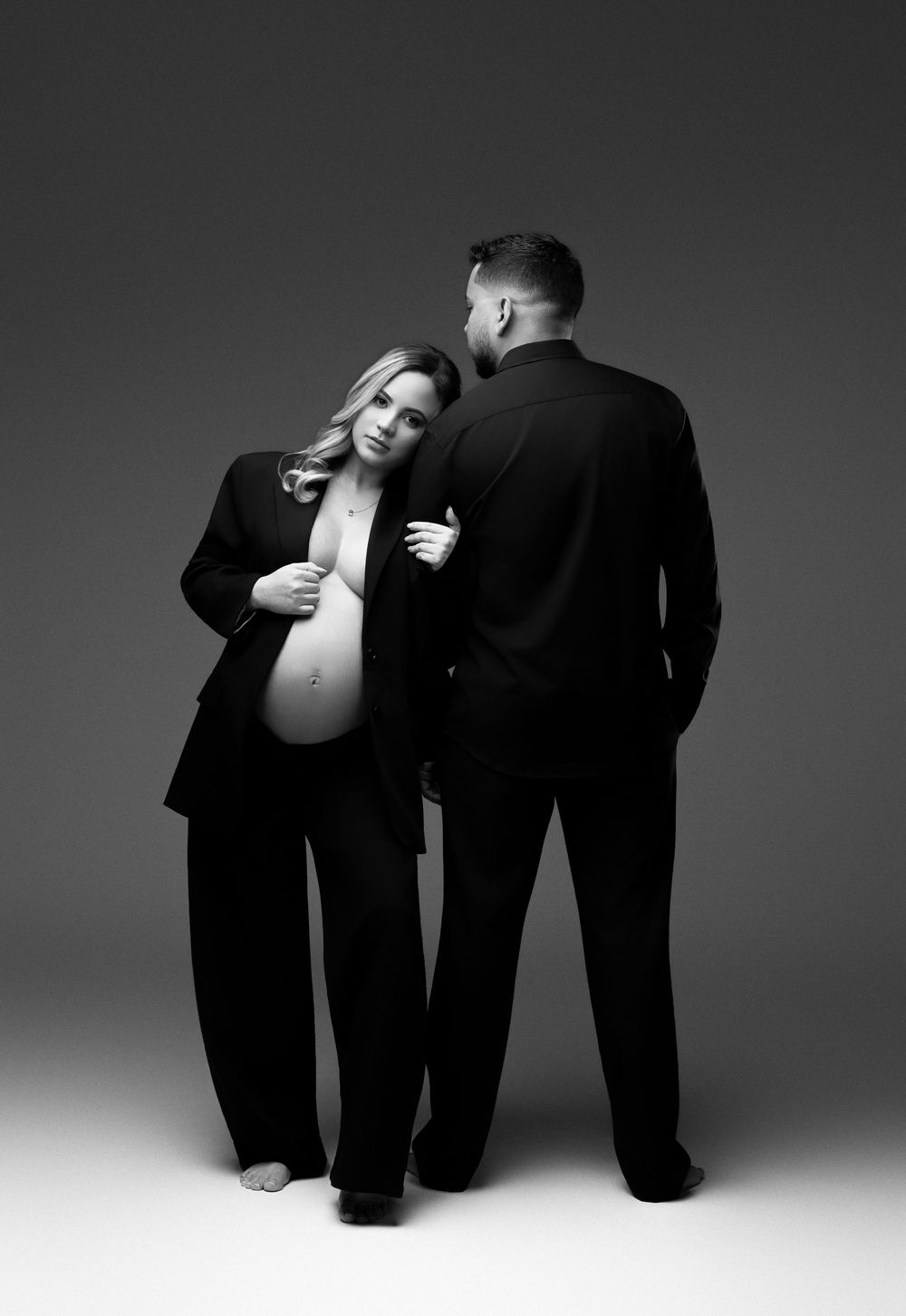 Black and white maternity photo of pregnant woman in suit leaning on partner, studio portrait.
