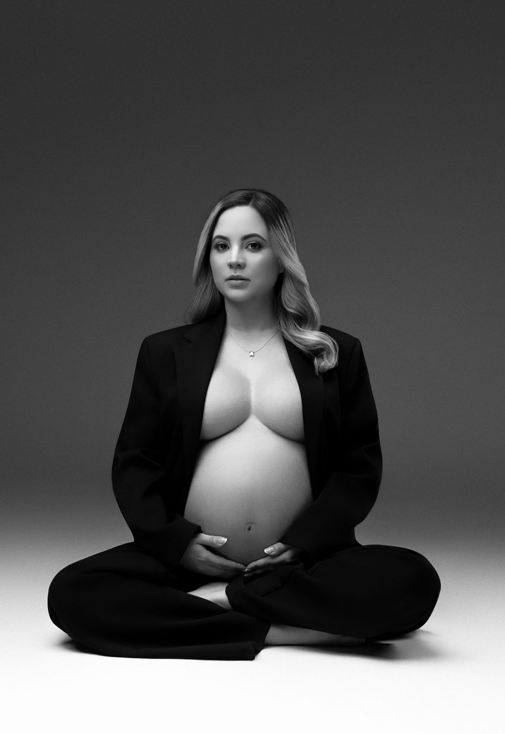 Black and white maternity photo of pregnant woman in open blazer sitting cross-legged on studio floor.