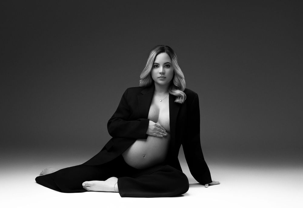 Black and white maternity photo of a blonde woman in an open blazer sitting gracefully on a studio floor.