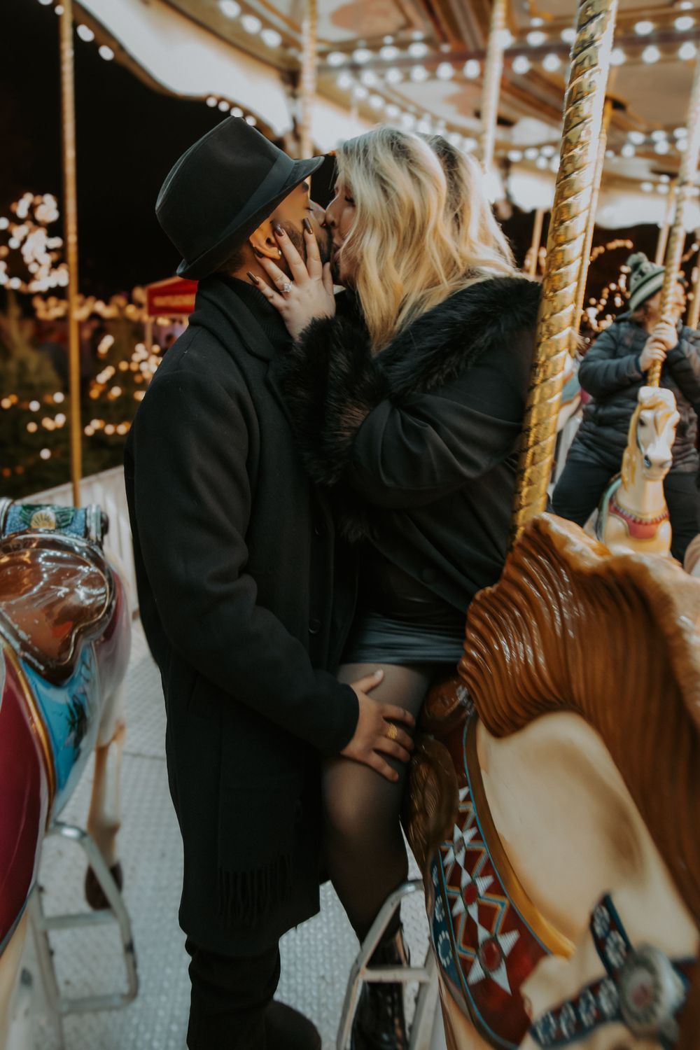 Couple in dark winter coats and a hat share a kiss beside a colorful carousel horse at a holiday market lit with twinkle lights.