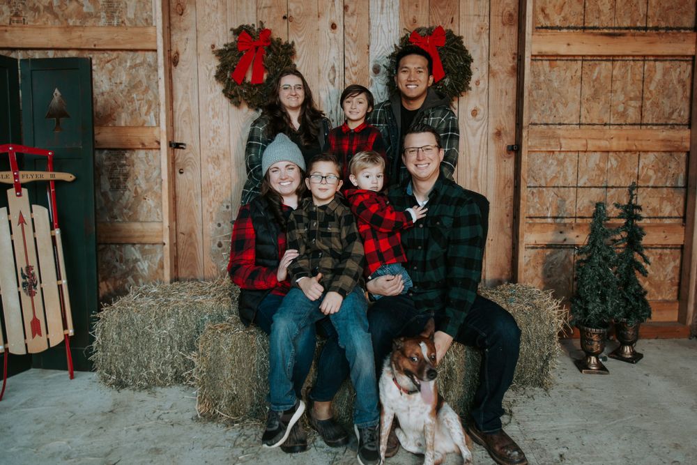 Family in plaid flannel shirts poses on hay bales with their dog beside red-bow Christmas wreaths on a rustic barn wall.