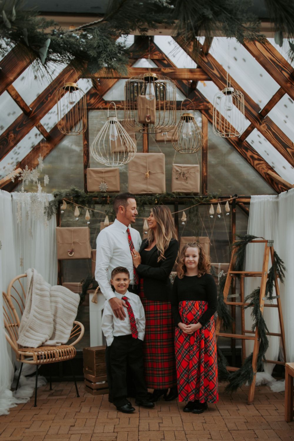 Family of four in red plaid and white shirts poses for a Christmas portrait in a rustic greenhouse with garland and hanging pendant lights.