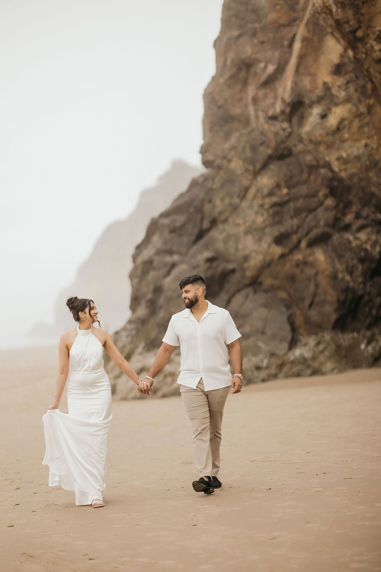 A couple in white and beige walk hand in hand along a foggy beach with cliffs in the background.