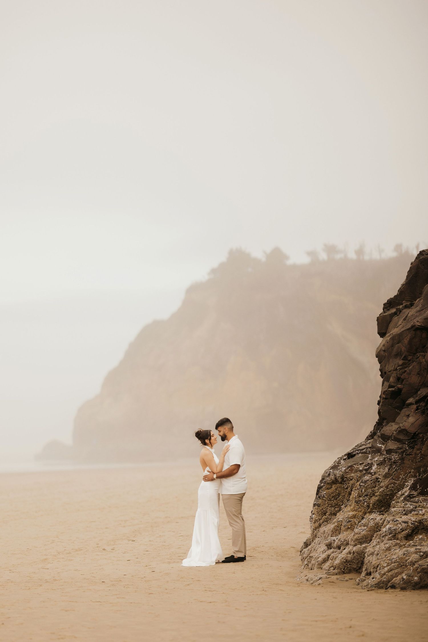 A couple embraces on a misty beach near dramatic coastal cliffs.
