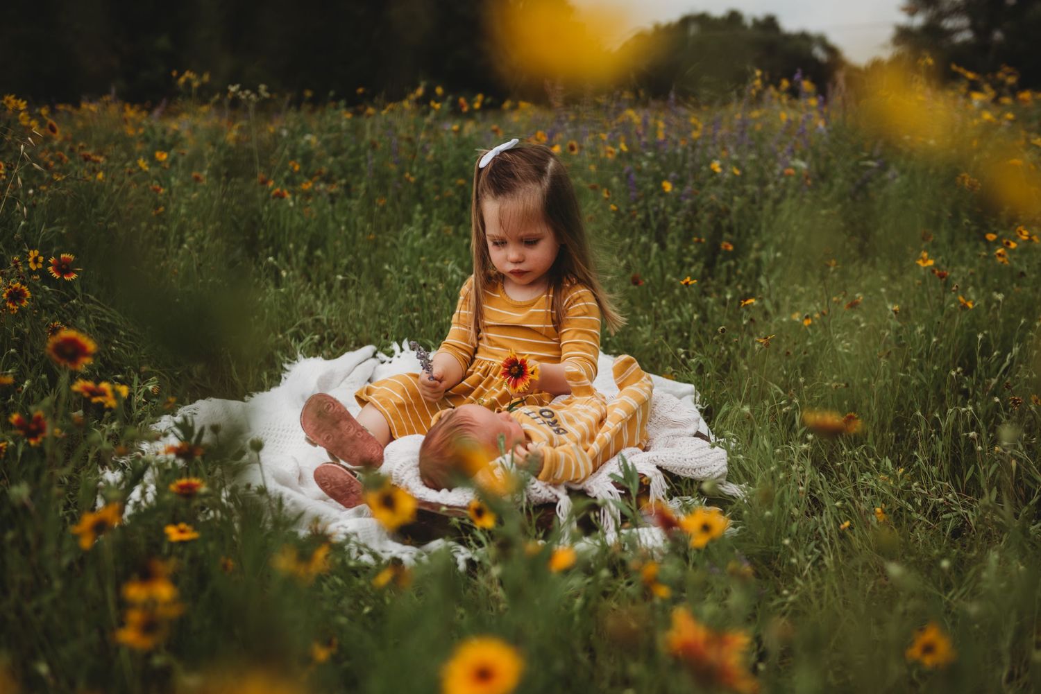 A small figure in a mustard dress sits on a white blanket surrounded by vibrant wildflowers in a summer meadow.