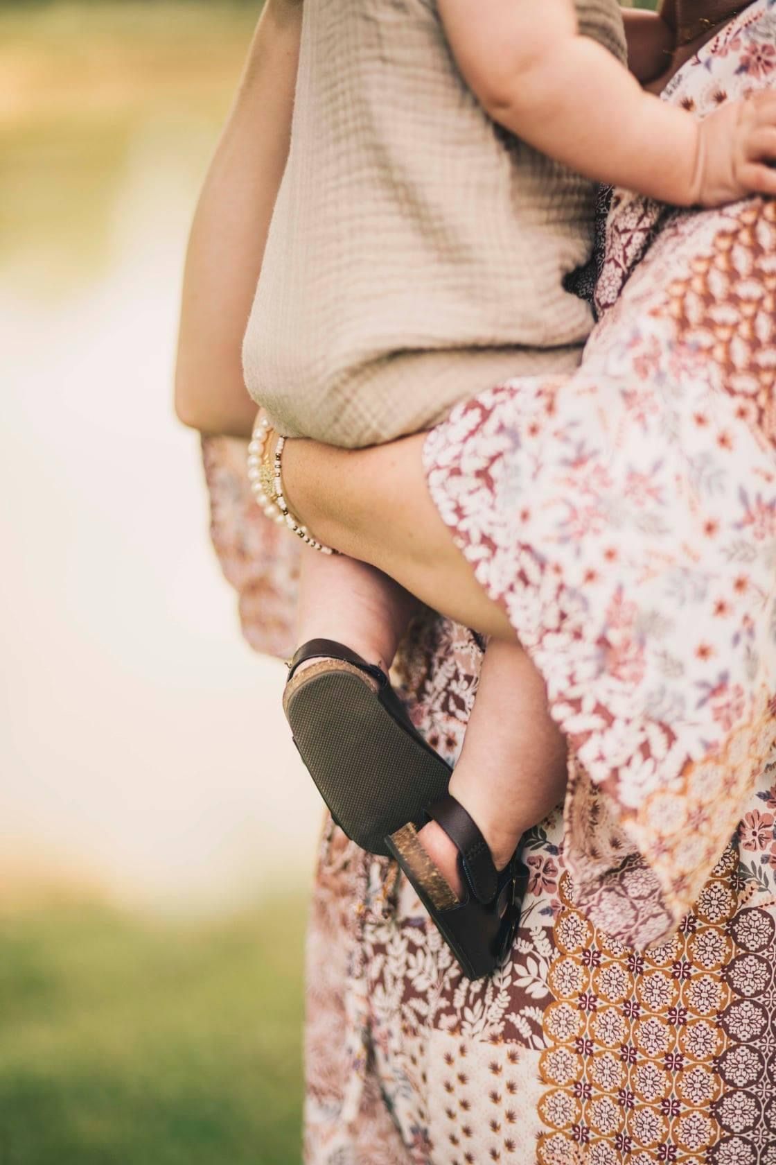 Close-up of a mother holding a baby wearing tiny black sandals, dressed in a floral skirt outdoors.