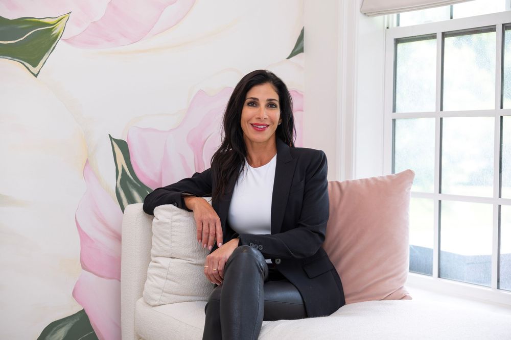 Professional woman in black blazer sits confidently on white sofa against floral wall, smiling warmly.