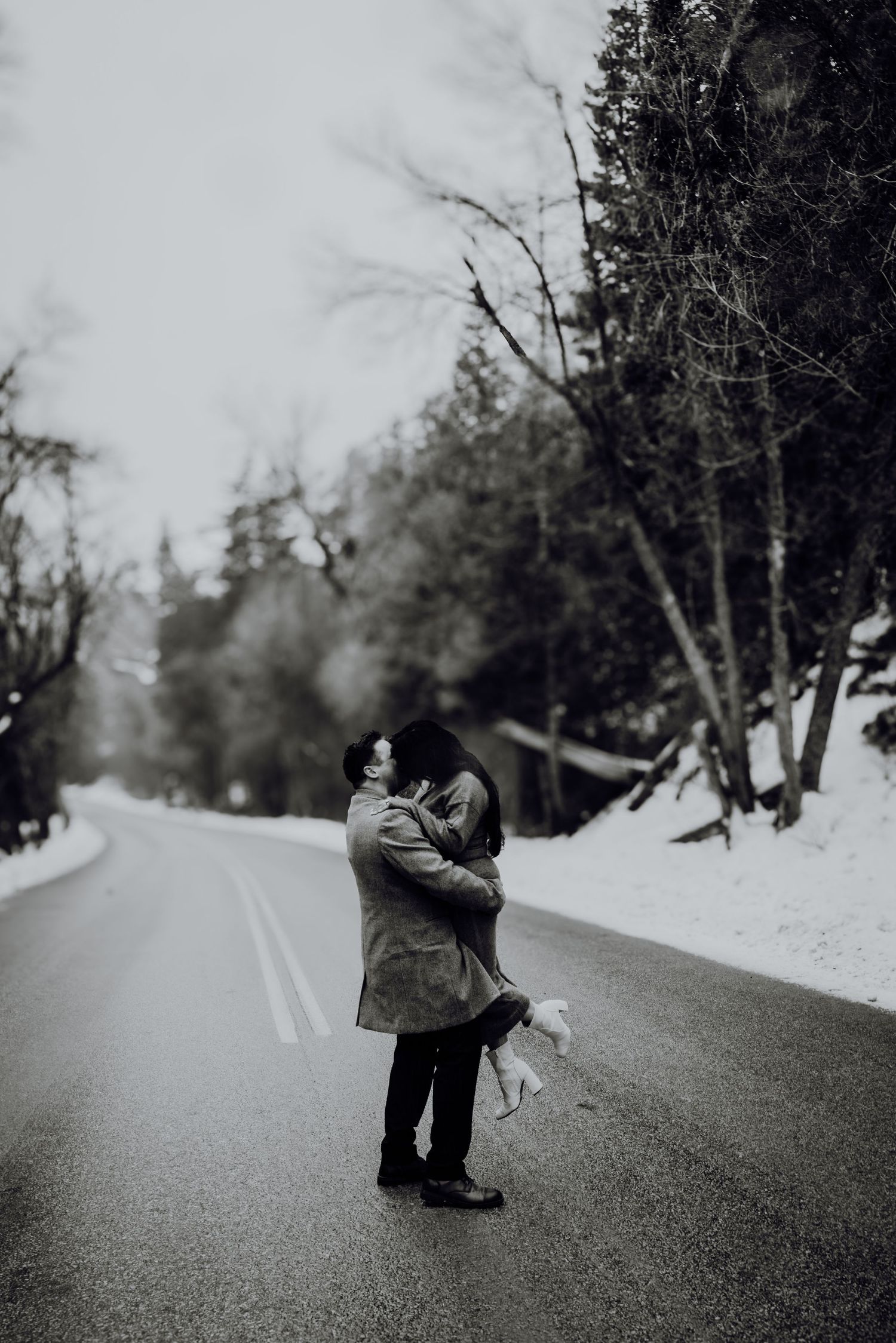 Couple embracing and kissing on a snowy winter road surrounded by bare trees in a romantic black and white photo.