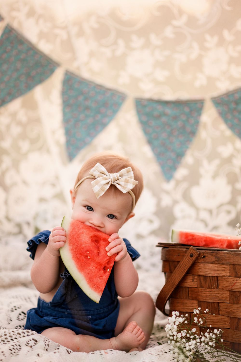 Adorable baby girl eating a watermelon slice beside a picnic basket with bunting banner backdrop.