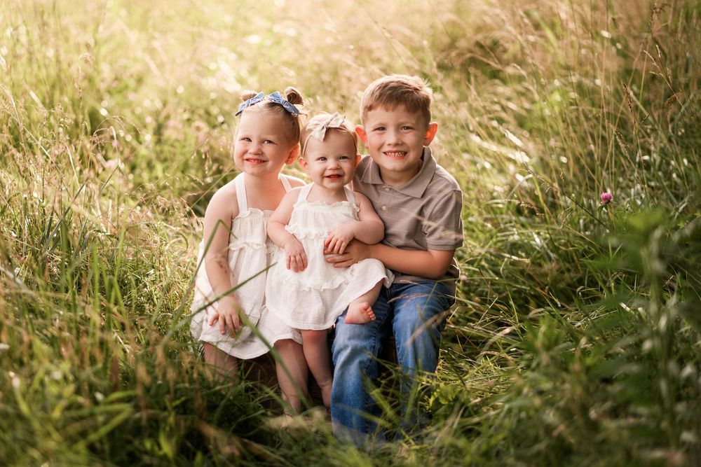 Three young siblings smiling together in a sunny meadow, two girls and a boy posing among tall green grass.