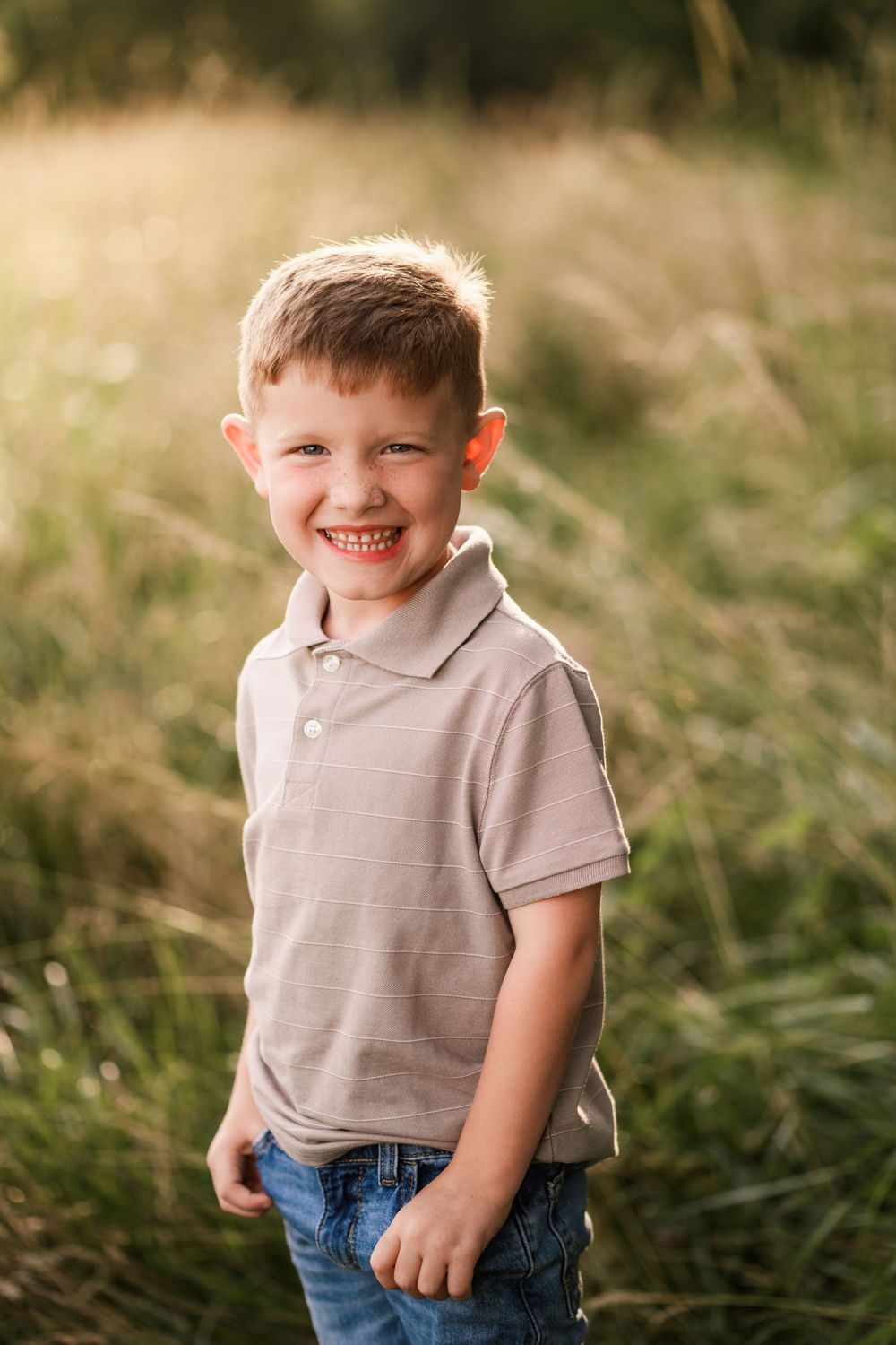 Smiling young boy in striped polo shirt standing in a sunlit green field during golden hour.