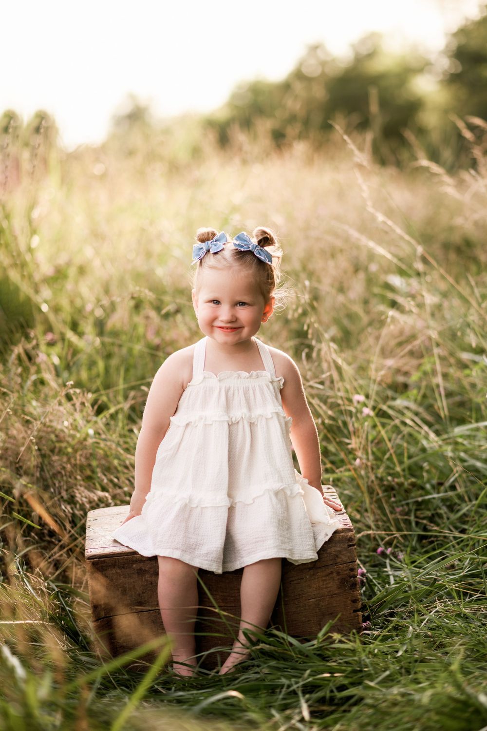 Smiling toddler girl in white dress sitting on wooden crate in sunny meadow with blue bow in hair.