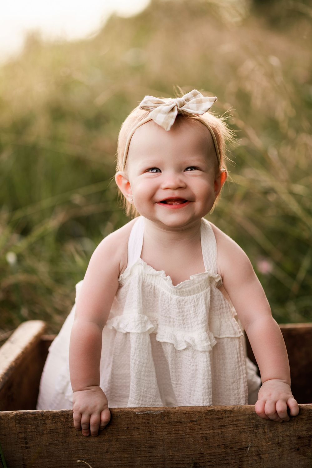 Happy baby girl in white ruffle dress with bow headband sitting in wooden crate in a sunny field.