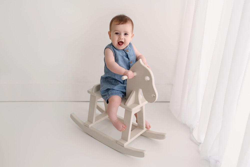 Baby in blue romper sits on a wooden rocking horse toy, smiling, in a bright white studio setting.