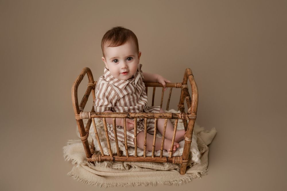 Baby sitting in a small rattan doll crib on a beige backdrop, wearing a striped romper.